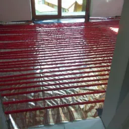 Czerwone rurki ogrzewania podłogowego ułożone na folii izolacyjnej z siatką, widok z góry na przygotowany system przed zalaniem wylewką.