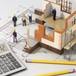 Model domu w trakcie budowy z cegły i pustaków, otoczony rusztowaniami, leżący na planie architektonicznym z miniaturowymi postaciami robotników, kalkulatorem i ołówkami.