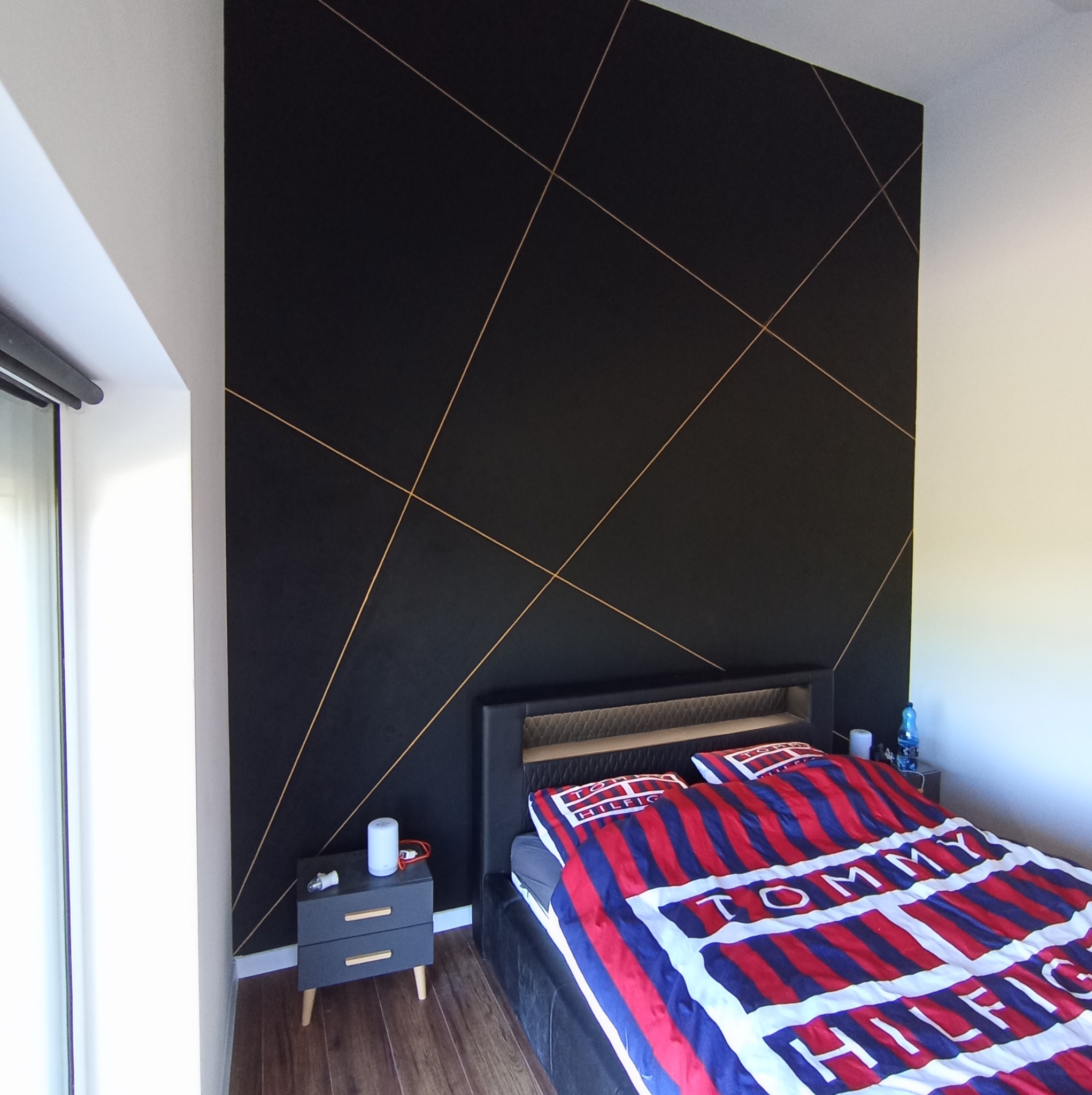 Sypialnia z czarną ścianą i złotymi geometrycznymi liniami, łóżko z pościelą Tommy Hilfiger, szara szafka nocna z drewnianymi nóżkami.