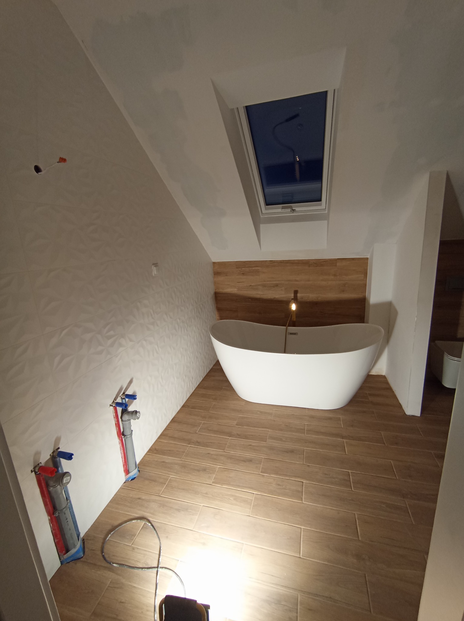 Nowoczesna łazienka na poddaszu z wolnostojącą białą wanną, drewnopodobnymi płytkami na podłodze i ścianie, białą ścianą z trójwymiarową strukturą, oknem dachowym i widocznymi elementami instalacji...