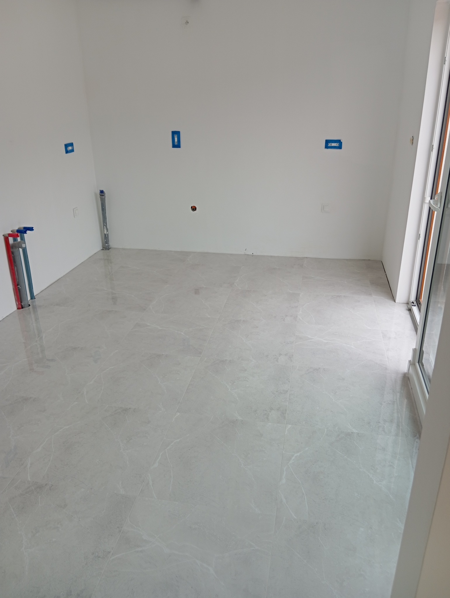 Pusty pokój z położonymi jasnoszarymi płytkami podłogowymi imitującymi marmur, przygotowane instalacje elektryczne i hydrauliczne, ściany pomalowane na biało.