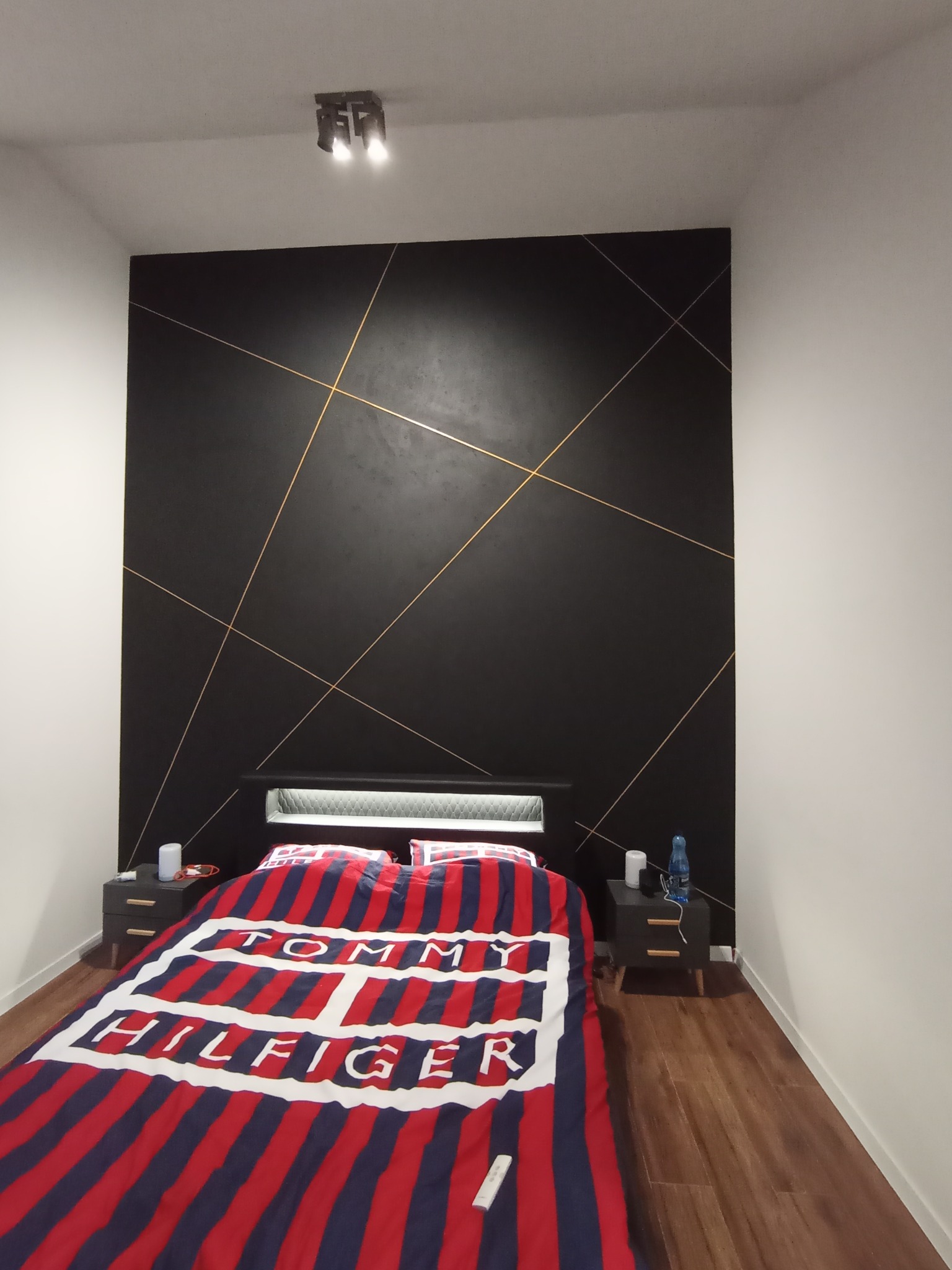 Sypialnia w Warszawie z czarną ścianą za łóżkiem, zdobioną złotymi liniami, oświetlona lampą sufitową, z drewnianą podłogą i szafkami nocnymi.