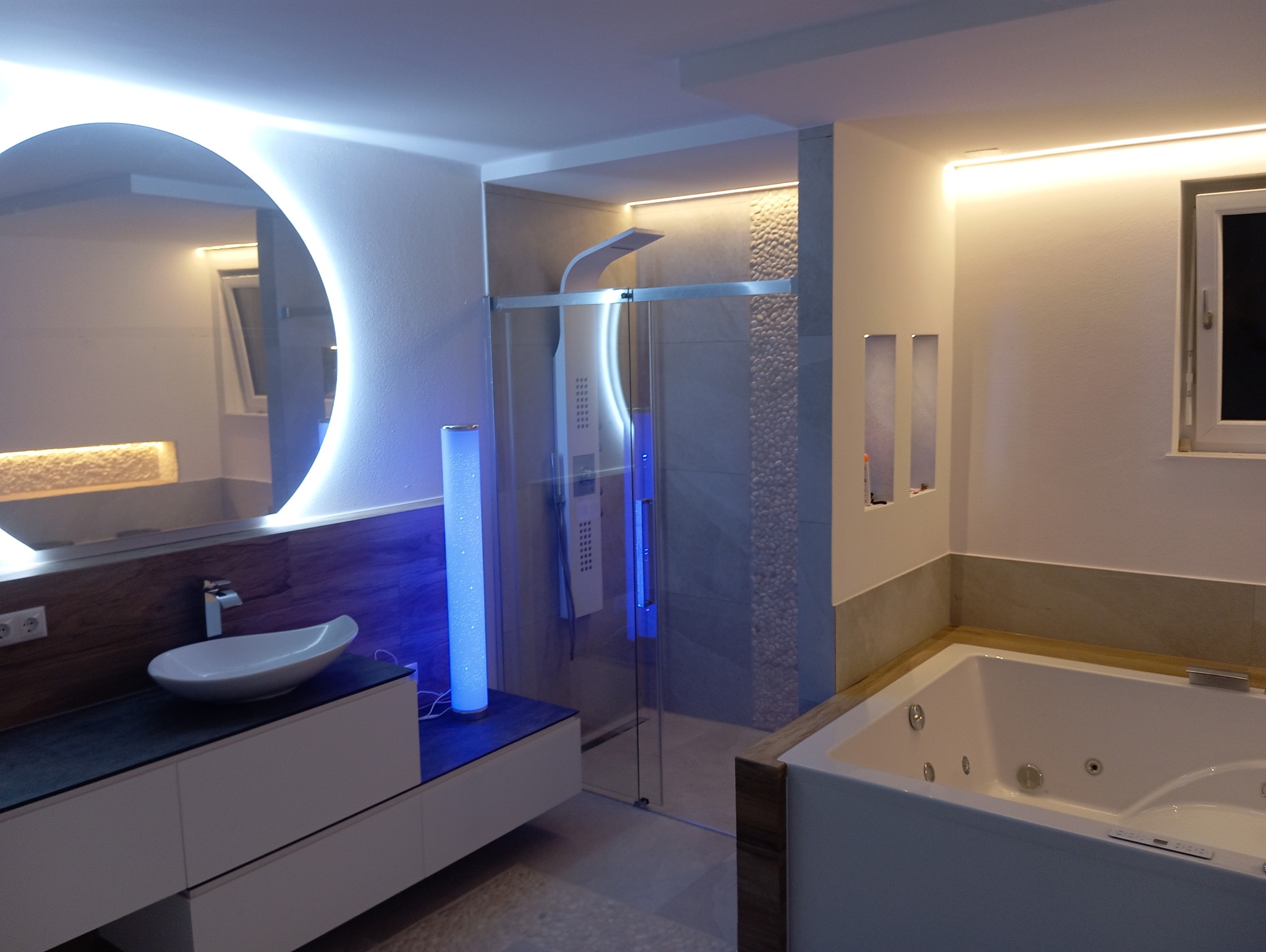 Nowoczesna łazienka z okrągłym lustrem podświetlanym LED, kabiną prysznicową ze szklanymi drzwiami i hydromasażem w wannie.