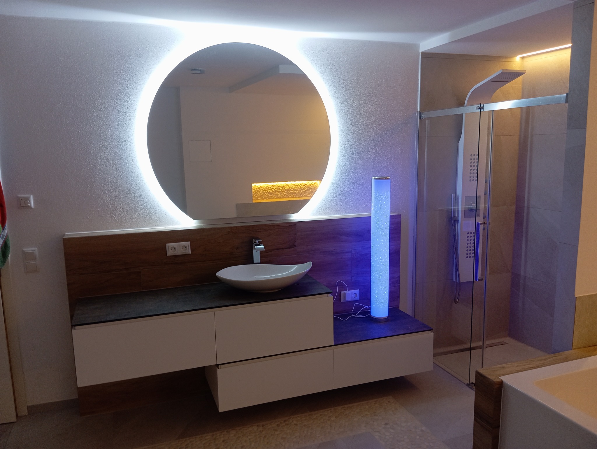 Nowoczesna łazienka z okrągłym lustrem podświetlanym LED, umywalką nablatową, szafką z szufladami, kabiną prysznicową typu walk-in i lampą podłogową z niebieskim światłem.