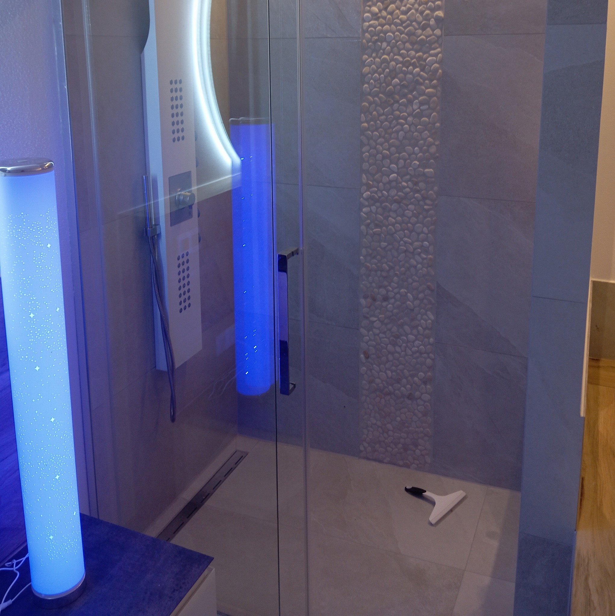 Nowoczesna kabina prysznicowa z oświetleniem LED, wykończona płytkami imitującymi kamień, z widoczną odpływem liniowym i ściągaczką do wody na podłodze.