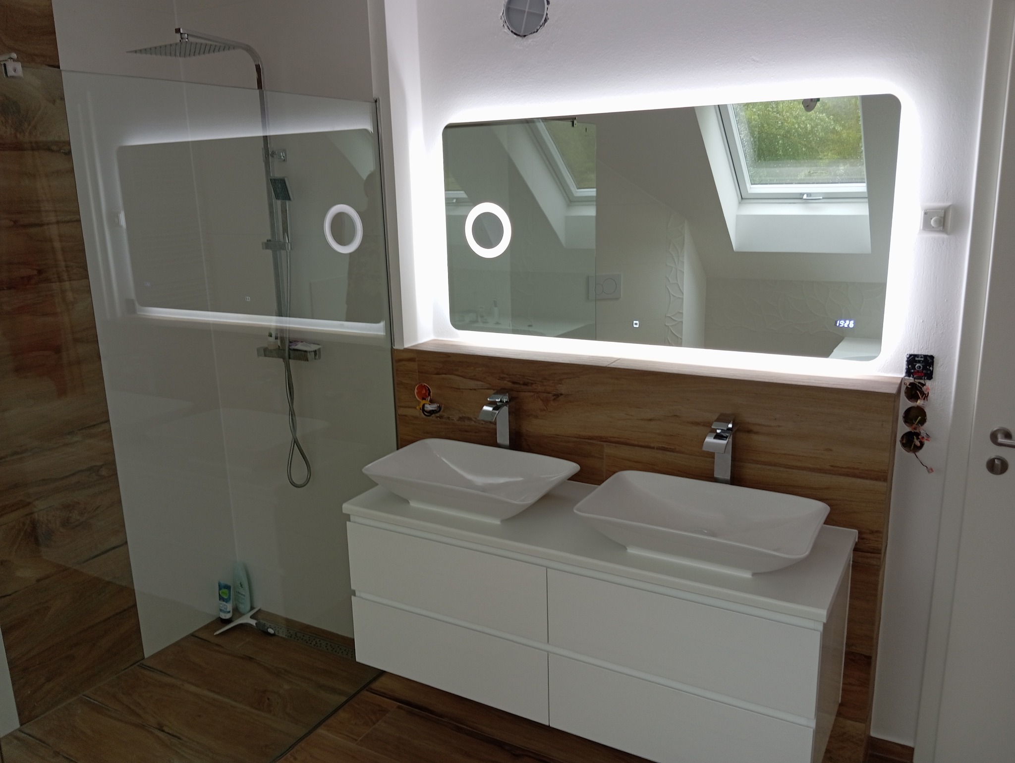 Nowoczesna łazienka z podwójną umywalką nablatową, podświetlanym lustrem z widoczną datą i prysznicem typu walk-in, wykończona drewnopodobnymi panelami na ścianie.