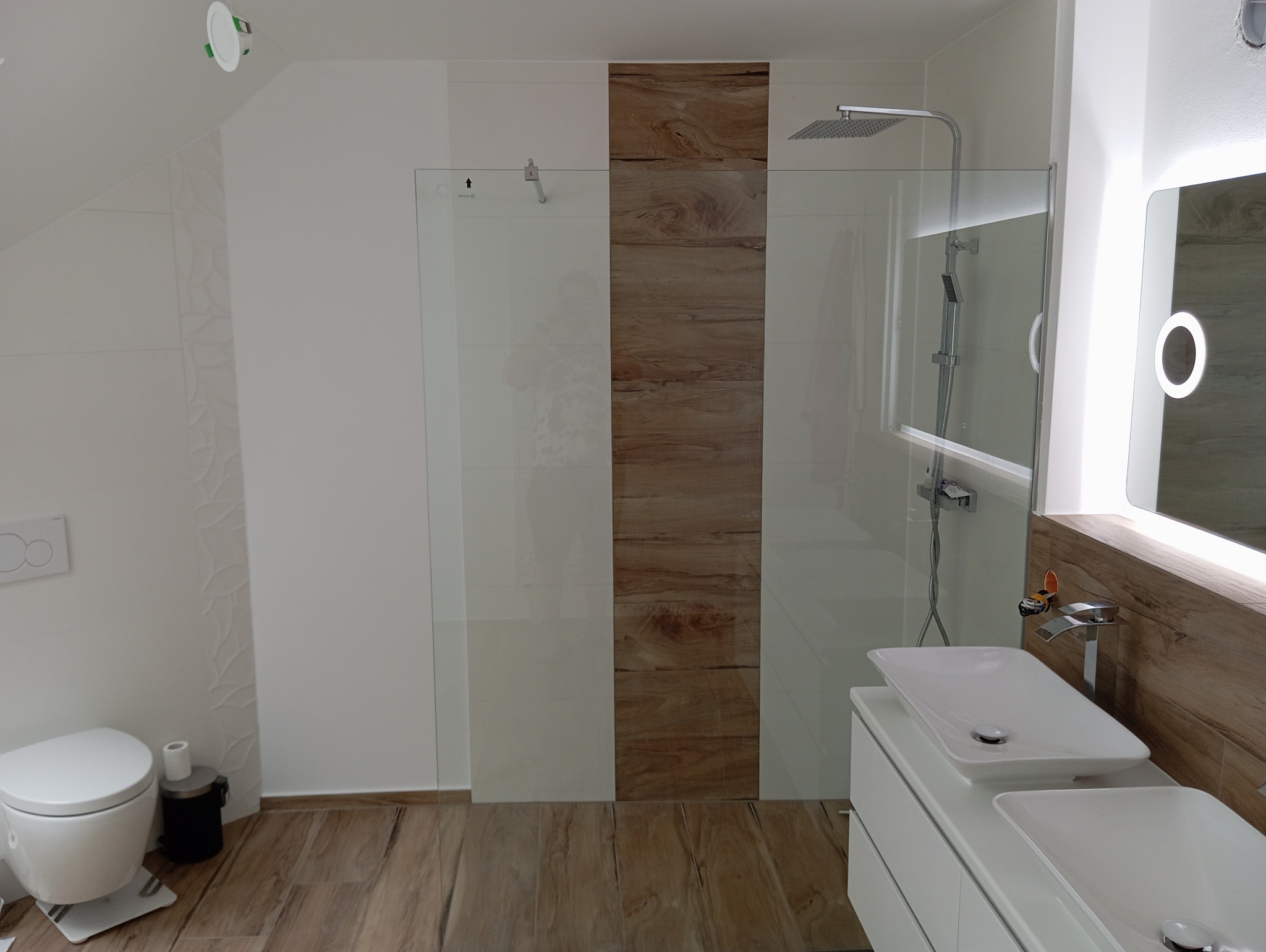 Nowoczesna łazienka z kabiną prysznicową typu walk-in, podwójną umywalką nablatową na białej szafce i podświetlanym lustrem, wykończona płytkami imitującymi drewno.