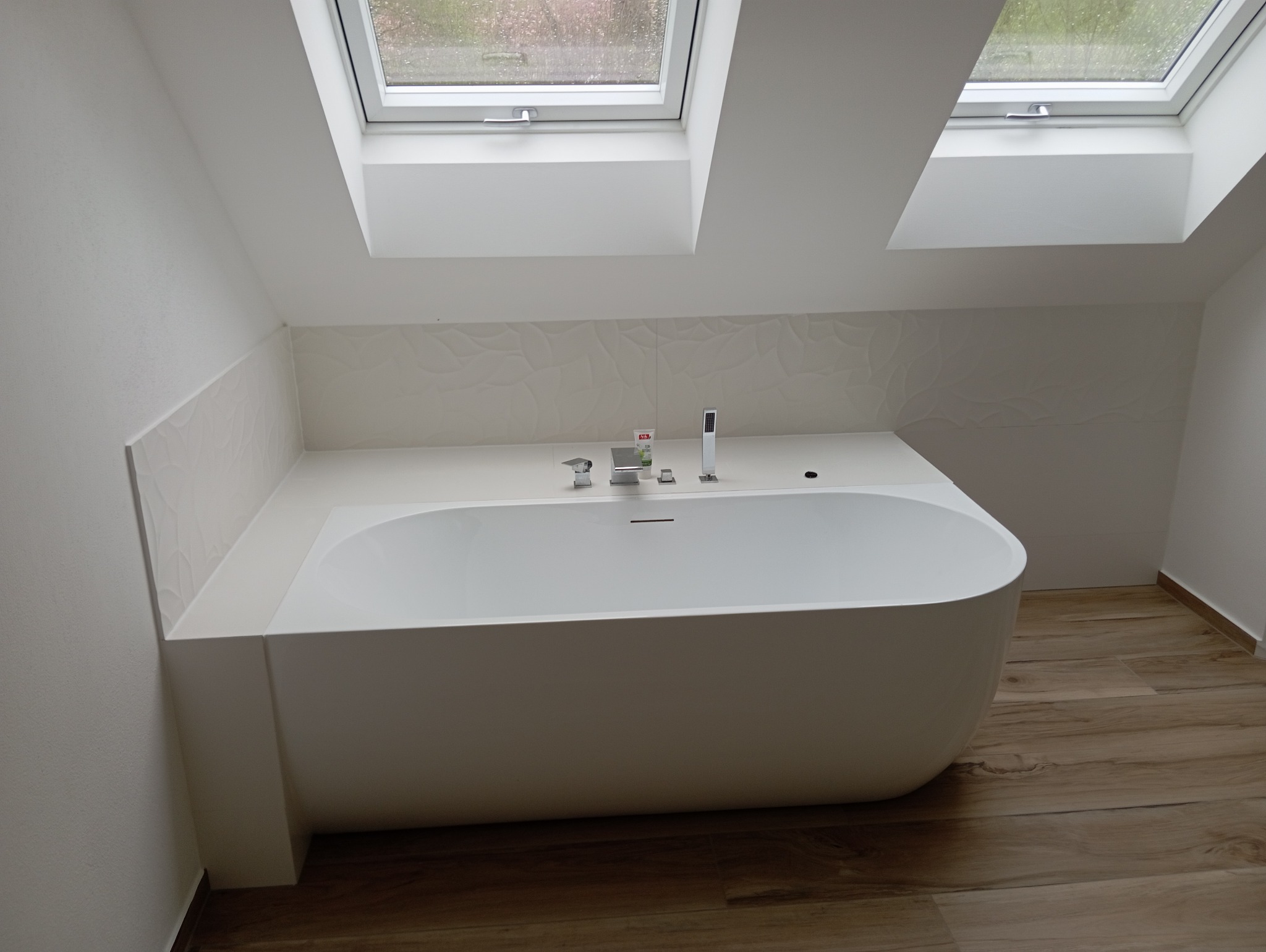 Nowoczesna, wolnostojąca wanna w łazience na poddaszu z dwoma oknami dachowymi, białe ściany z dekoracyjną fakturą płytek, armatura łazienkowa ze srebrnymi bateriami i kosmetykami.