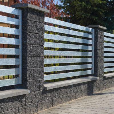 Nowoczesne ogrodzenie z szarych, łupanych bloczków betonowych i poziomych, metalowych przęseł, widoczne z bliska na tle zieleni drzew.
