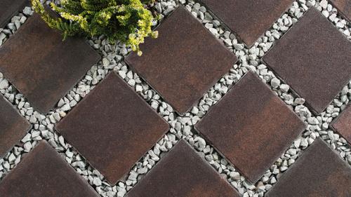 Ułożona nawierzchnia z kwadratowych, brązowych płyt, wypełniona drobnymi, szarymi kamieniami, z fragmentem zielonej rośliny w lewym górnym rogu.