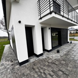 zloterolety.pl - Nowoczesny dom z białymi ścianami i czarnymi akcentami: bramy garażowe, okna, balkon z czarną balustradą. Kostka brukowa w odcieniach szarości.