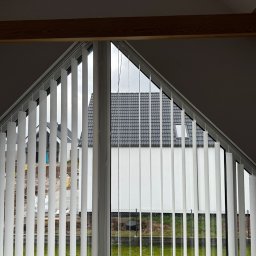 zloterolety.pl - Białe żaluzje pionowe zamontowane w oknie na poddaszu, widok na dom z szarą dachówką i zielony trawnik. Ujęcie z wnętrza, z widocznymi elementami konstrukcji dachu.