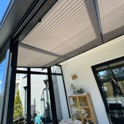 zloterolety.pl - Beżowe plisy dachowe zamontowane w nowoczesnej, przeszklonej werandzie z czarnymi ramami okiennymi. Widok z dołu na harmonijkowo złożone plisy.