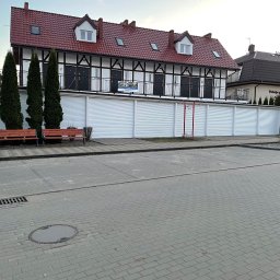 zloterolety.pl - Budynek z czerwonym dachem i białymi roletami garażowymi. Widoczne drzewa, ławki i kostka brukowa. Ujęcie z poziomu ulicy, w tle budynek 'Pokoje gościnne'.