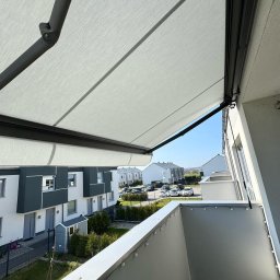 zloterolety.pl - Balkon z rozłożoną, jasną markizą w nowoczesnym budownictwie. Widok z góry na osiedle domów i błękitne niebo. Oświetlenie słoneczne.