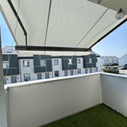 zloterolety.pl - Balkon z rozłożoną, jasną markizą przeciwsłoneczną, sztuczną trawą i dekoracyjnymi lampkami. W tle widok na nowoczesne budynki mieszkalne w słoneczny dzień.