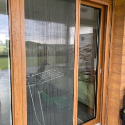 zloterolety.pl - Zamknięta moskitiera plisowana w drewnianej ramie okiennej, widok z bliska. W tle widoczny ogród i suszarka na pranie. Ochrona przed owadami w nowoczesnym domu.