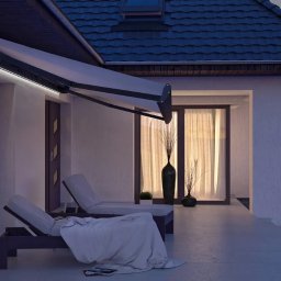 zloterolety.pl - Elegancka, rozłożona markiza tarasowa z oświetleniem LED przy białym domu z dwoma leżakami na tarasie, widok wieczorny.