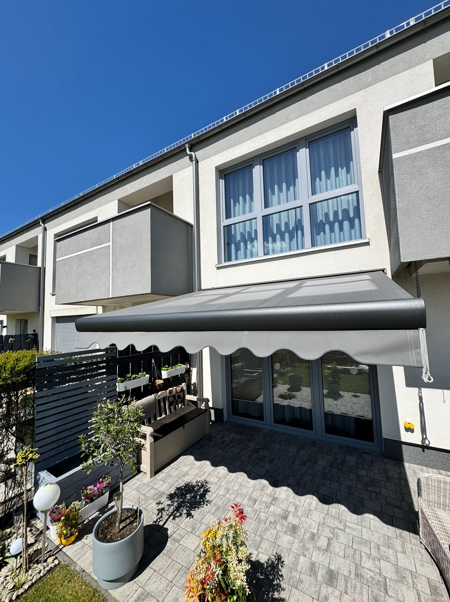 Szara markiza tarasowa z falbaną, zamontowana nad przeszklonymi drzwiami w nowoczesnym domu z balkonami. Widok na patio z roślinami i meblami ogrodowymi w słoneczny dzień.