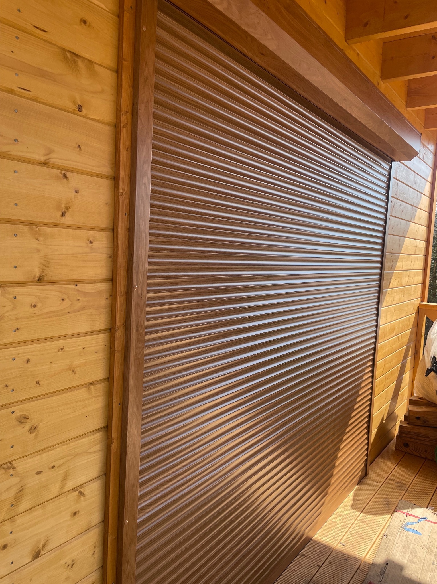 Zamknięta, brązowa roleta zewnętrzna na drewnianej elewacji budynku. Widoczne poziome lamele i prowadnice rolety. Ujęcie z dołu, podkreślające strukturę drewna.