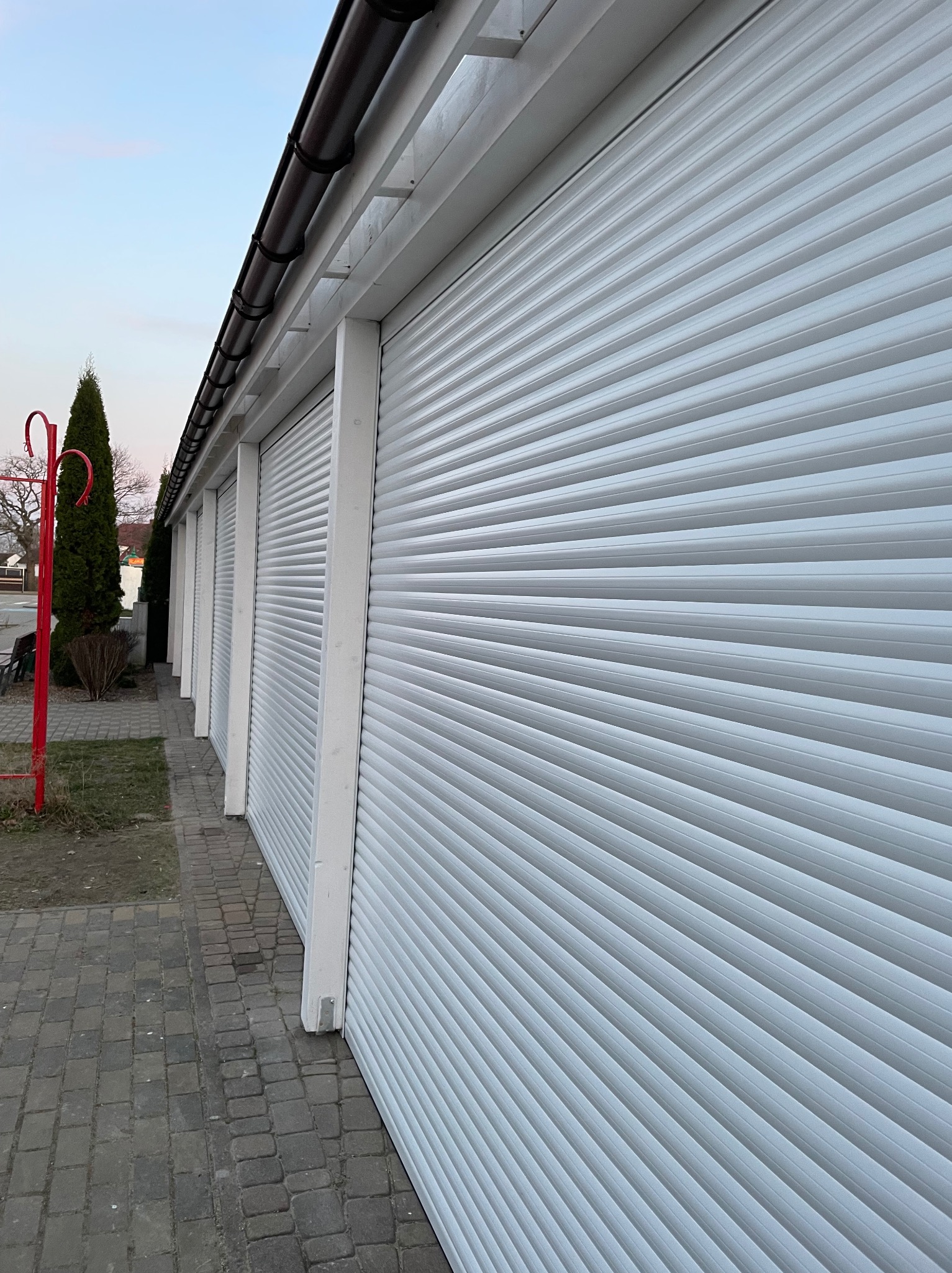 Białe rolety zewnętrzne na budynku, widok z boku, z perspektywą zbieżną. Widoczny fragment chodnika z kostki brukowej i elementy zieleni. Ujęcie z dołu.