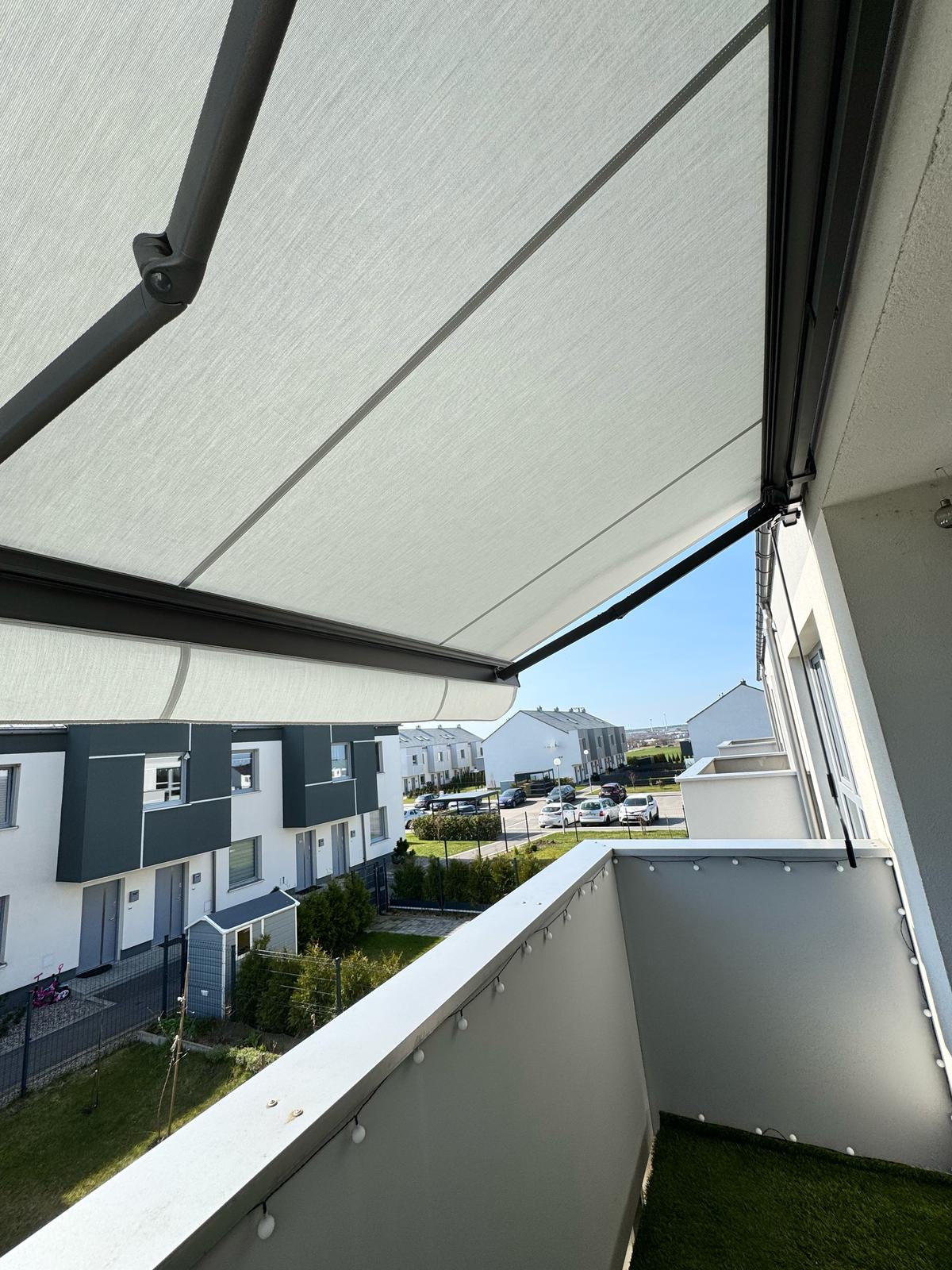 Balkon z rozłożoną, jasną markizą w nowoczesnym budownictwie. Widok z góry na osiedle domów i błękitne niebo. Oświetlenie słoneczne.