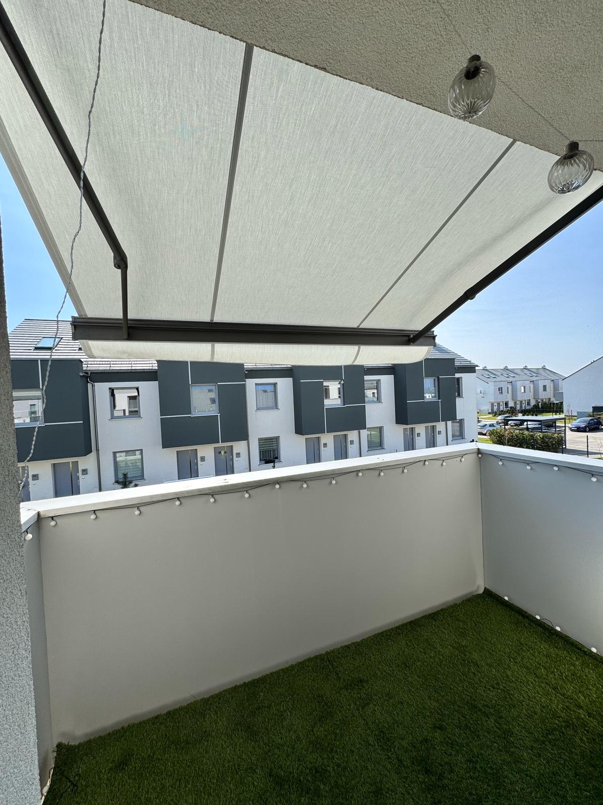 Balkon z rozłożoną, jasną markizą przeciwsłoneczną, sztuczną trawą i dekoracyjnymi lampkami. W tle widok na nowoczesne budynki mieszkalne w słoneczny dzień.