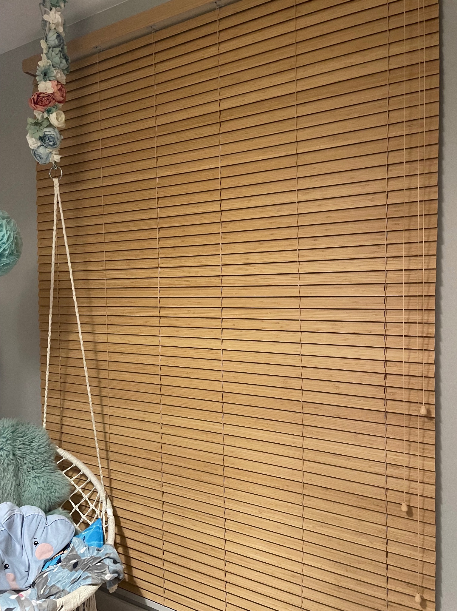 Jasne, drewniane żaluzje poziome zamontowane w pokoju dziecięcym, widoczne z boku huśtawki z poduszkami i dekoracją kwiatową.