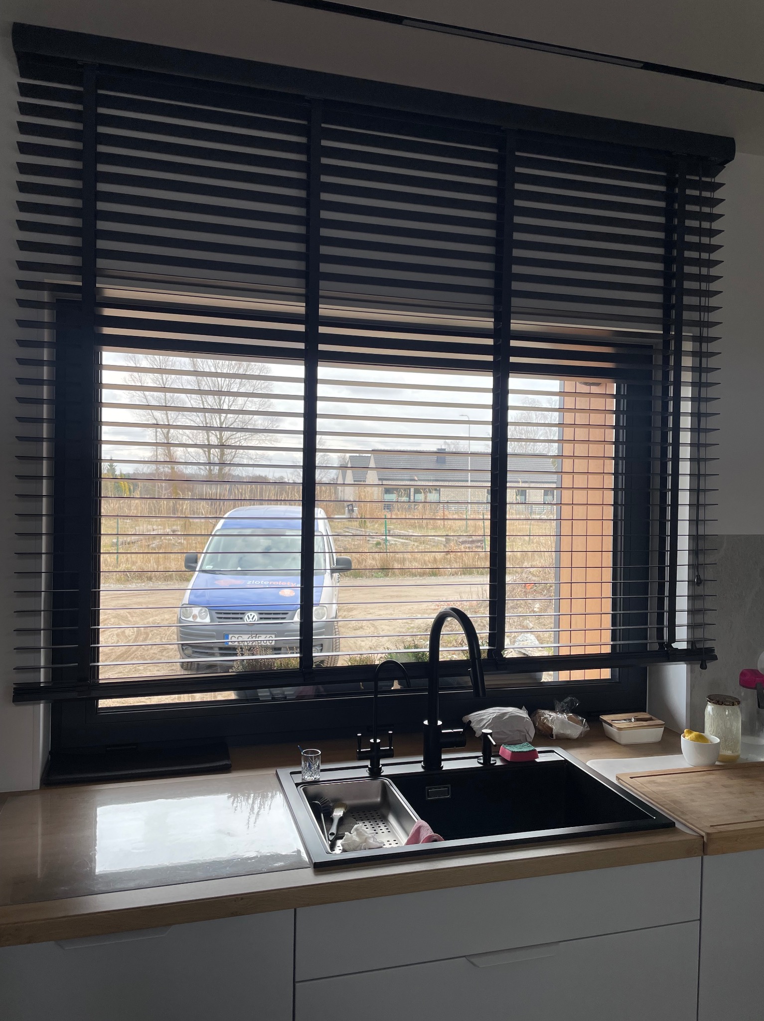 Ciemne żaluzje drewniane poziome w oknie kuchennym, widok na zewnątrz z zaparkowanym niebieskim busem. Minimalistyczny wystrój kuchni, nowoczesne wykończenie.