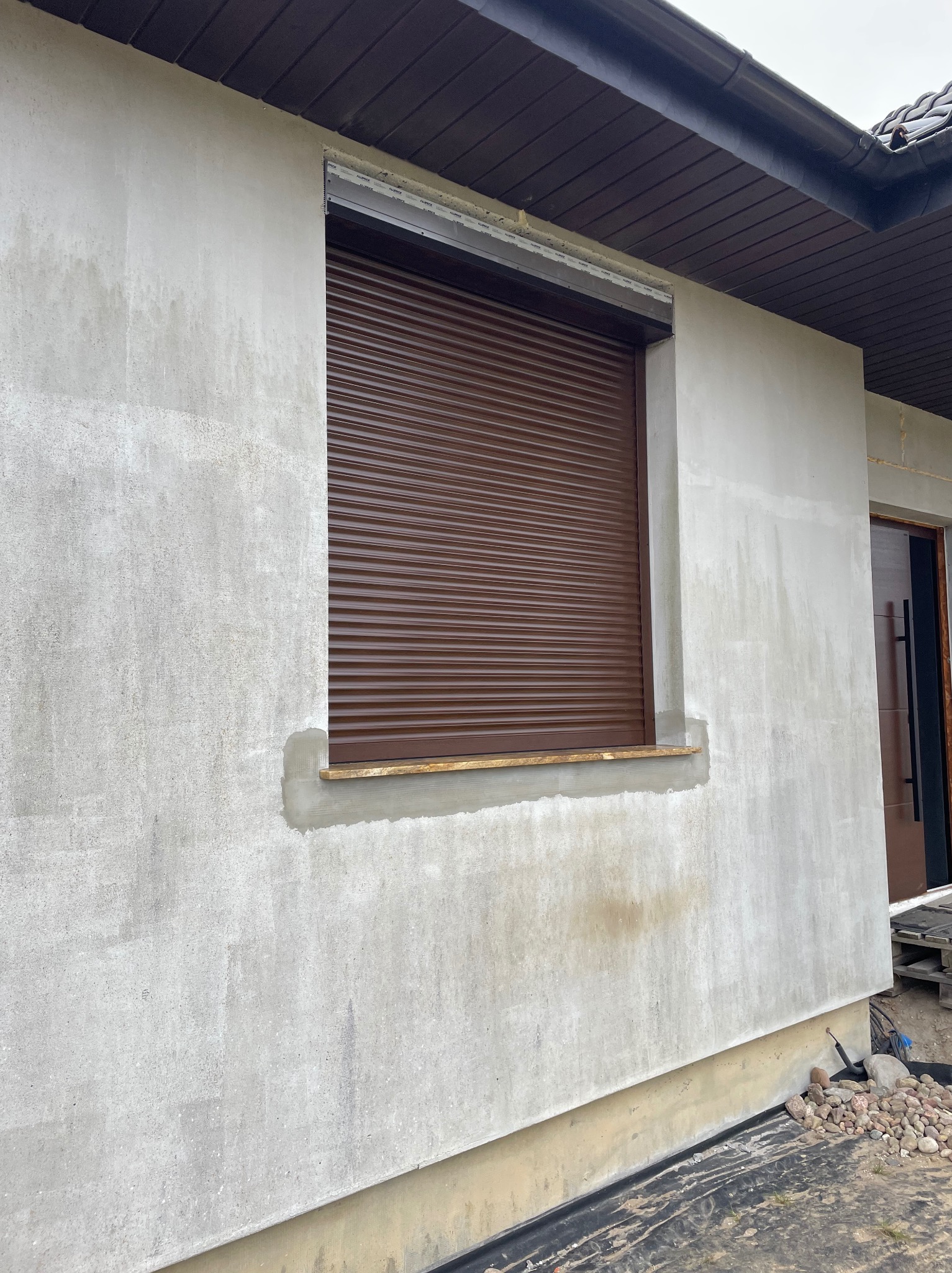 Brązowa roleta zewnętrzna zamontowana w oknie budynku w trakcie wykończenia. Widoczny surowy tynk i fragment dachu. Ujęcie z boku, z dołu.