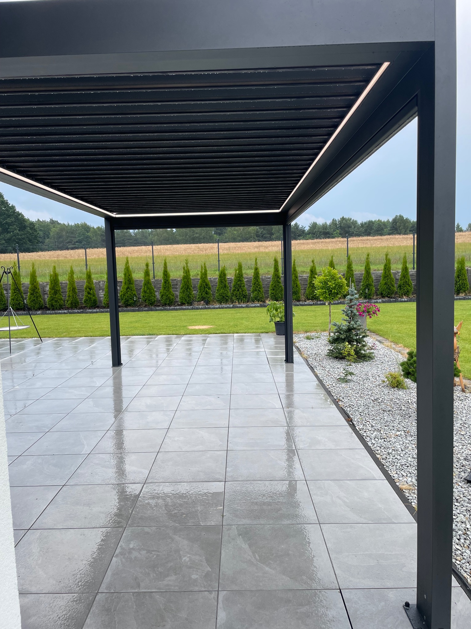 Nowoczesna, czarna pergola z oświetleniem LED na tarasie z szarych płyt. W tle zadbany trawnik i rząd tuj. Widok na ogród z elementami małej architektury.