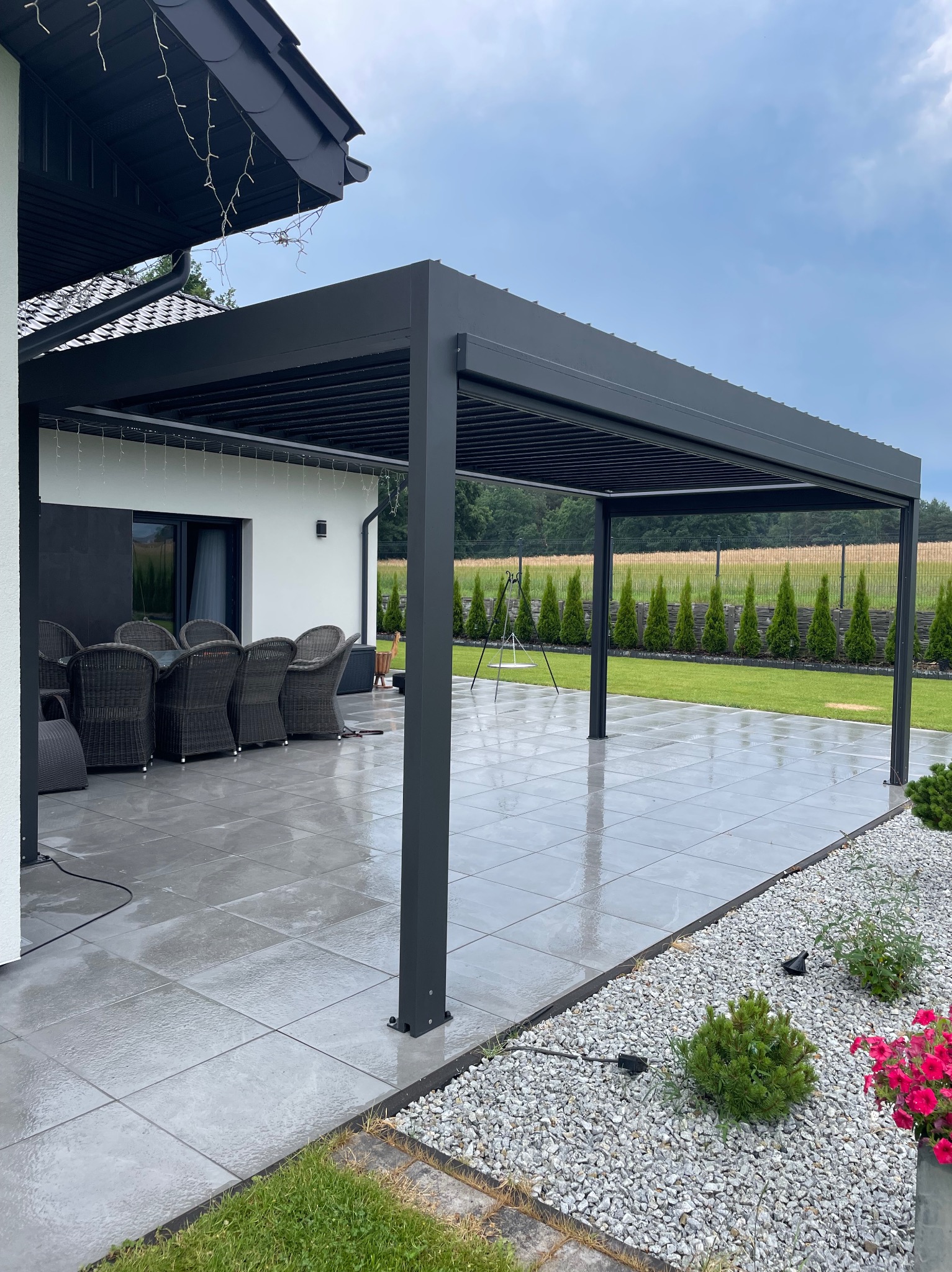 Nowoczesna, grafitowa pergola z lamelami na tarasie z szarych płyt. W tle zielony trawnik i rząd tuj. Widoczne meble ogrodowe i oświetlenie.