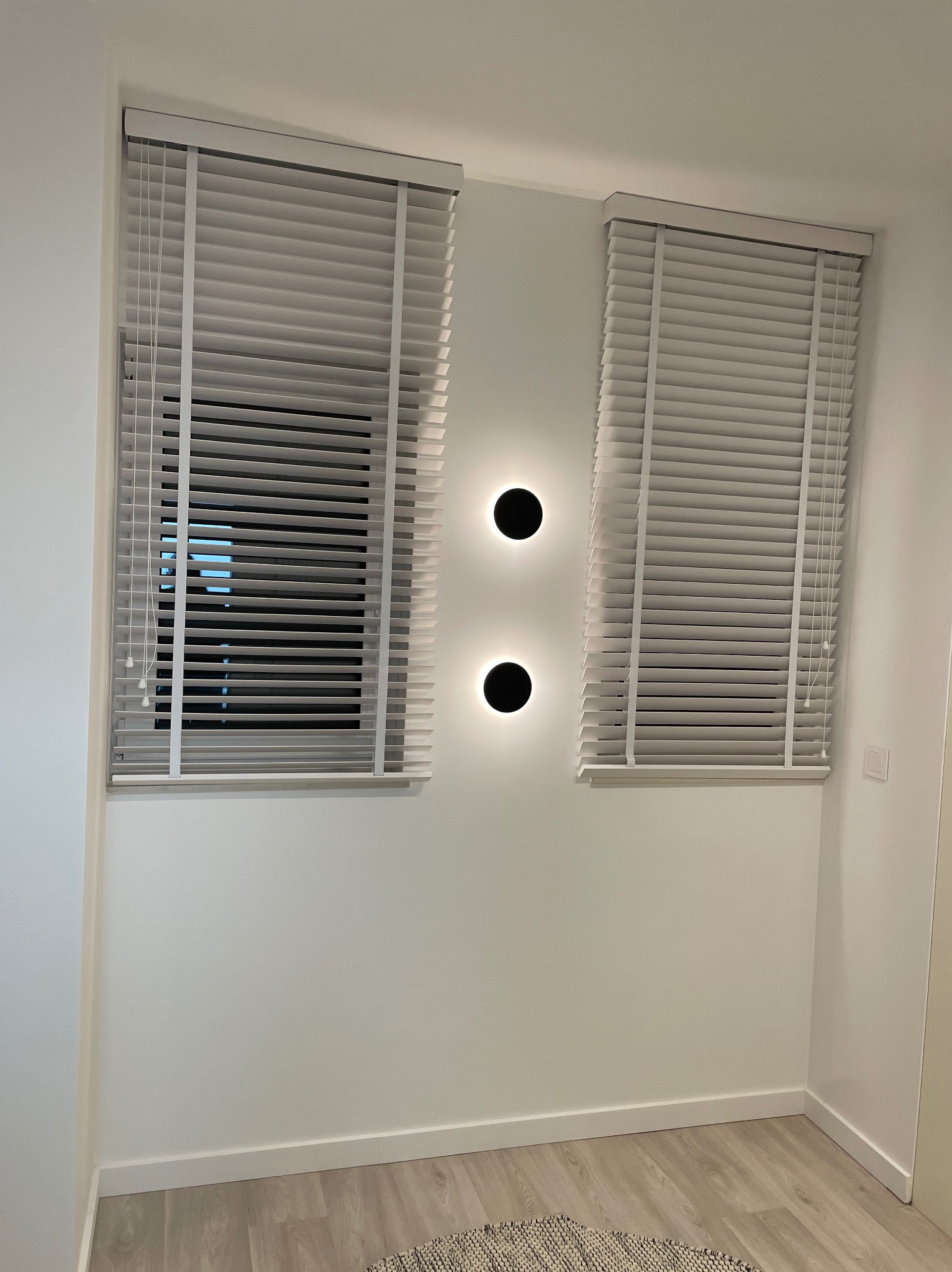 Dwa okna z białymi żaluzjami drewnianymi, częściowo otwarte, z czarnymi, okrągłymi lampami ściennymi pomiędzy nimi. Minimalistyczne wnętrze z jasną podłogą.