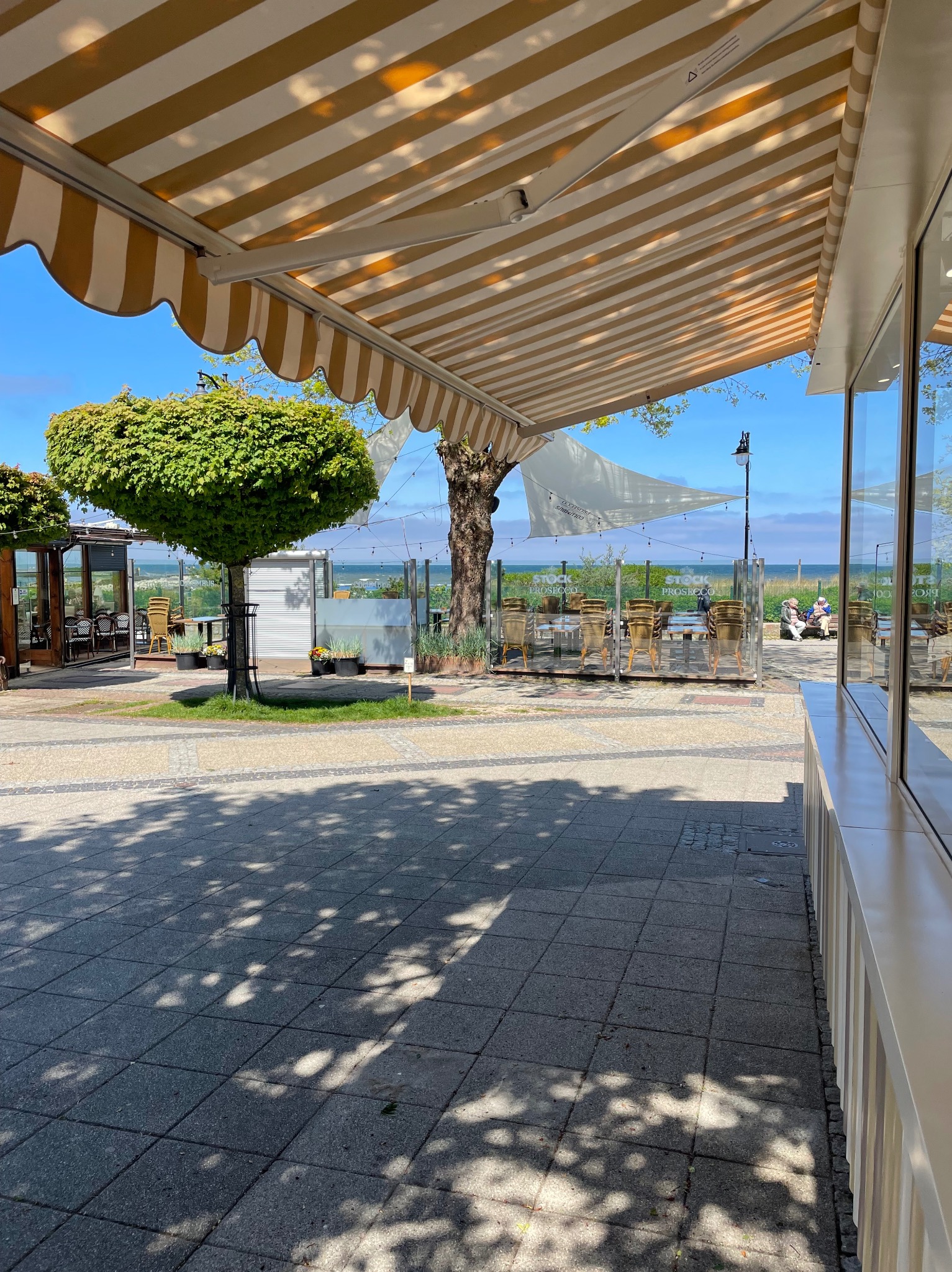 Beżowo-biała markiza nad letnim ogródkiem restauracji z widokiem na morze. Cień na chodniku, drzewa i stoliki w tle. Słoneczny dzień.