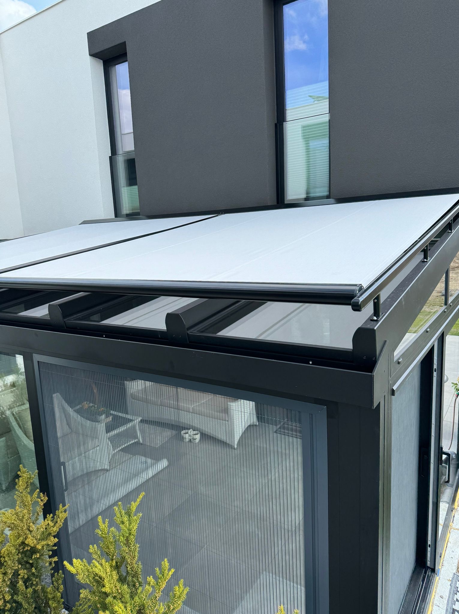 Nowoczesna pergola z rozsuwanym, jasnym zadaszeniem i pionową moskitierą w grafitowej ramie, na tle dwukondygnacyjnego domu z oknami.