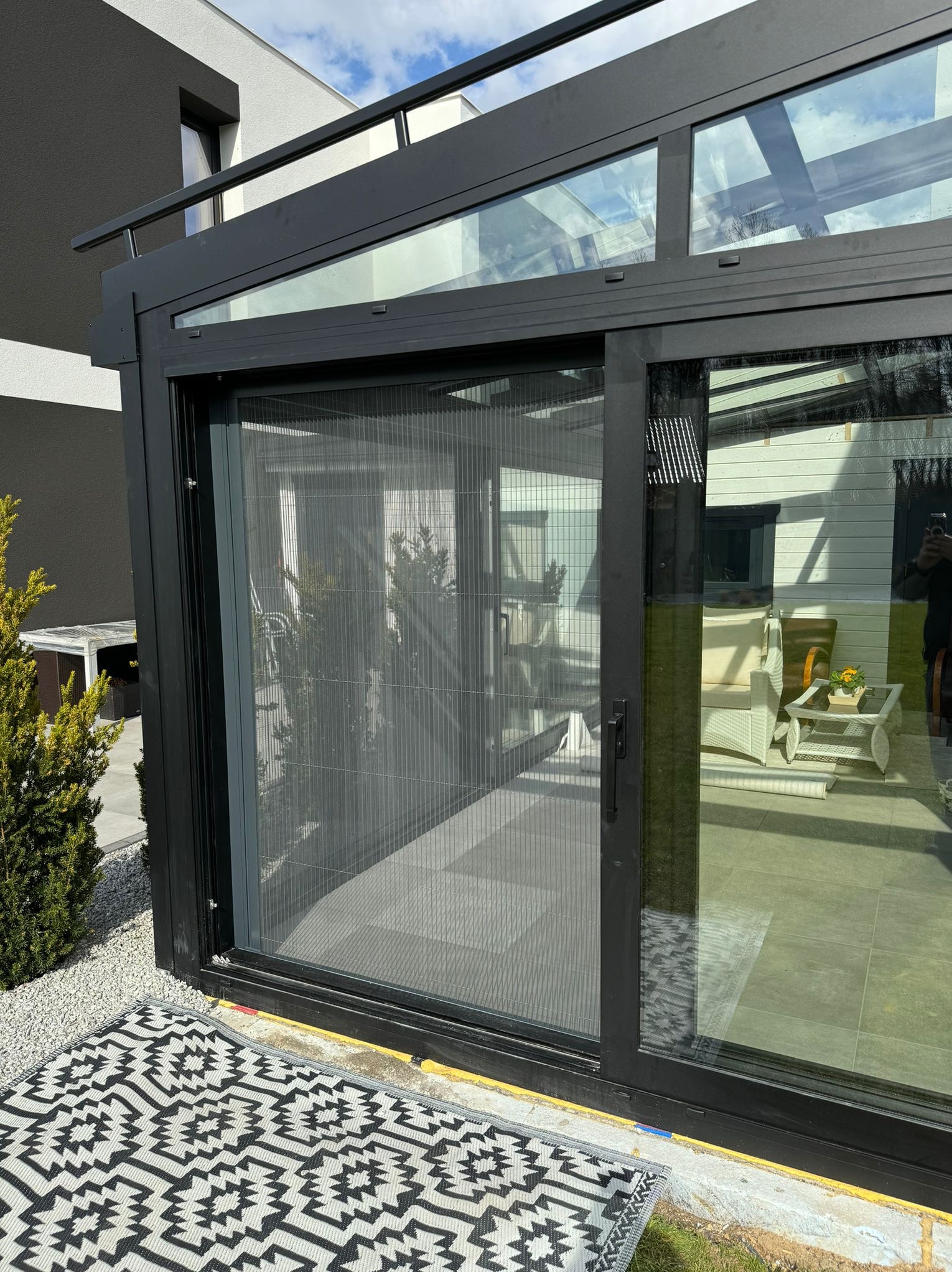 Nowoczesna, czarna rama okienna z siatką przeciw owadom, widok z zewnątrz na taras z meblami ogrodowymi. Dywanik z geometrycznym wzorem na pierwszym planie.