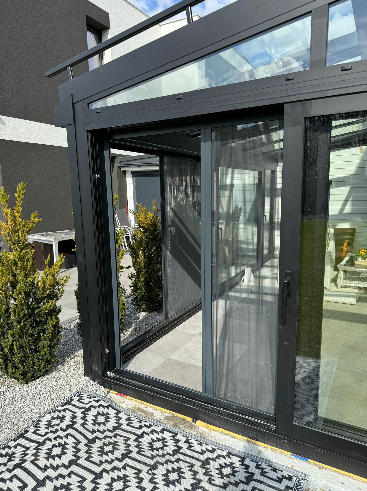 Nowoczesna zabudowa tarasowa z przesuwną moskitierą w kolorze antracytowym, widok z zewnątrz na taras z roślinami i dywanem w geometryczny wzór.