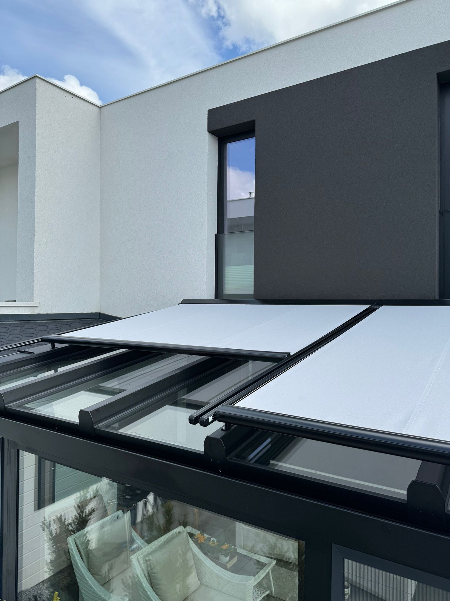 Nowoczesna pergola z rozsuwanym, białym zadaszeniem na tle dwukolorowej elewacji budynku. Widok z dołu, podkreślający detale konstrukcji i minimalistyczny design.