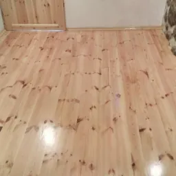 Jasne, drewniane deski podłogowe z widocznymi sękami, ułożone w pomieszczeniu z fragmentem kamiennej ściany i drzwiami z tego samego drewna.