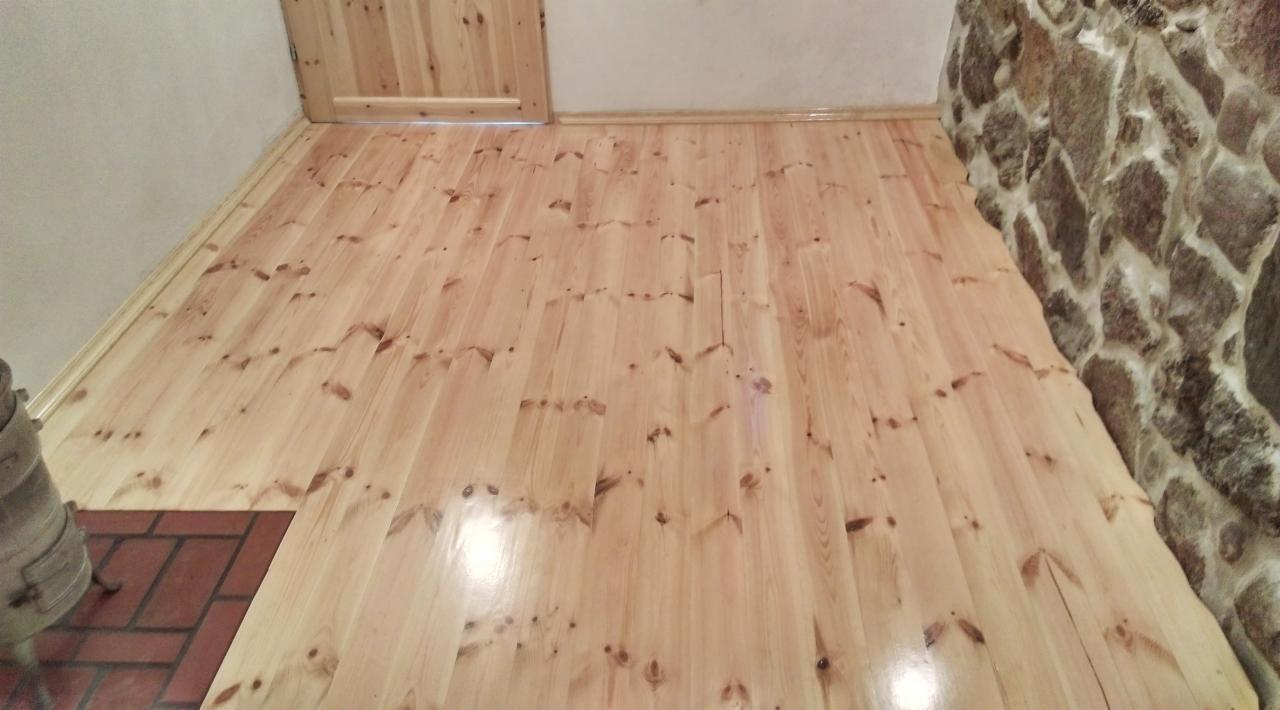 Jasne, drewniane deski podłogowe z widocznymi sękami, ułożone w pomieszczeniu z fragmentem kamiennej ściany i drzwiami z tego samego drewna.