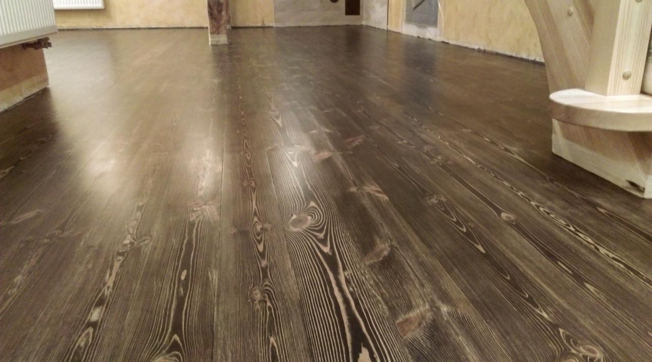 Ciemnobrązowa, lakierowana podłoga z desek sosnowych o wyraźnym usłojeniu, widziana pod kątem, z fragmentem jasnego, drewnianego elementu schodów w prawym górnym rogu.