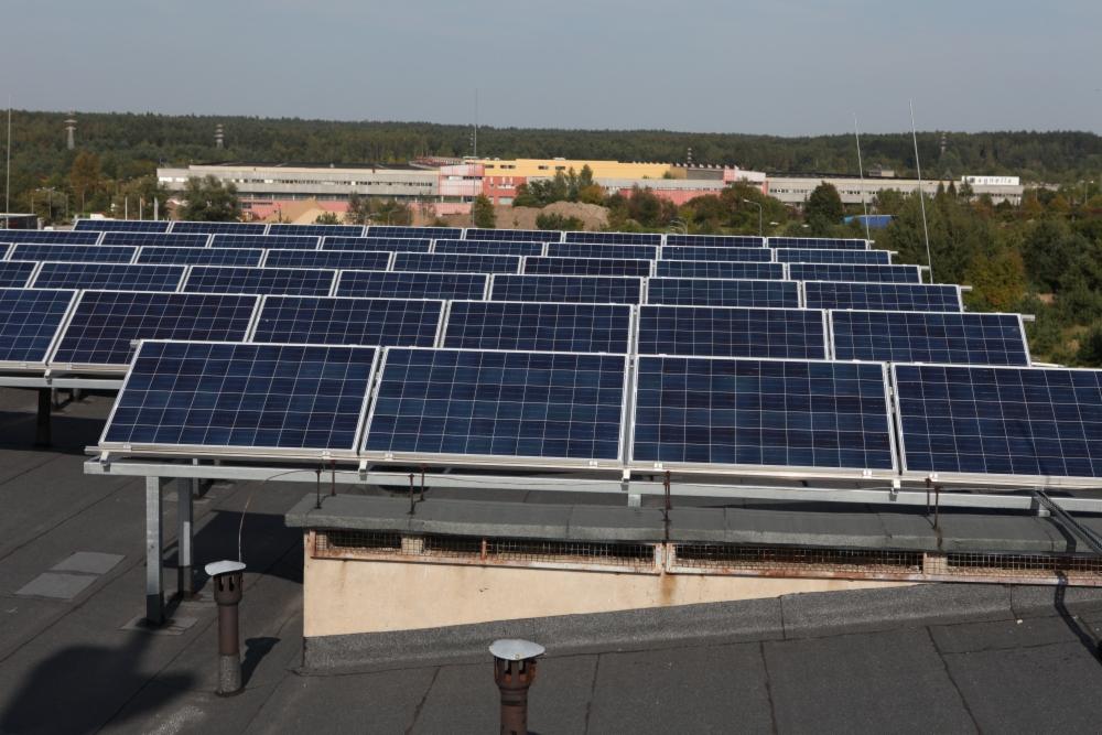 Rząd paneli słonecznych na płaskim dachu budynku przemysłowego, z widocznymi kominkami wentylacyjnymi i budynkami w tle.