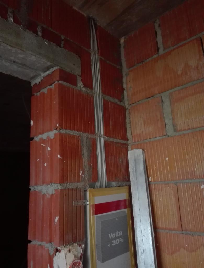 Pionowe kable elektryczne w bruzdach ceglanej ściany, widoczna puszka instalacyjna Volta +30% i aluminiowa listwa. Instalacja w trakcie wykonywania.
