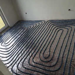 Niebieskie rury ogrzewania podłogowego ułożone w pętle na izolacji z siatką w surowym pomieszczeniu z oknem.