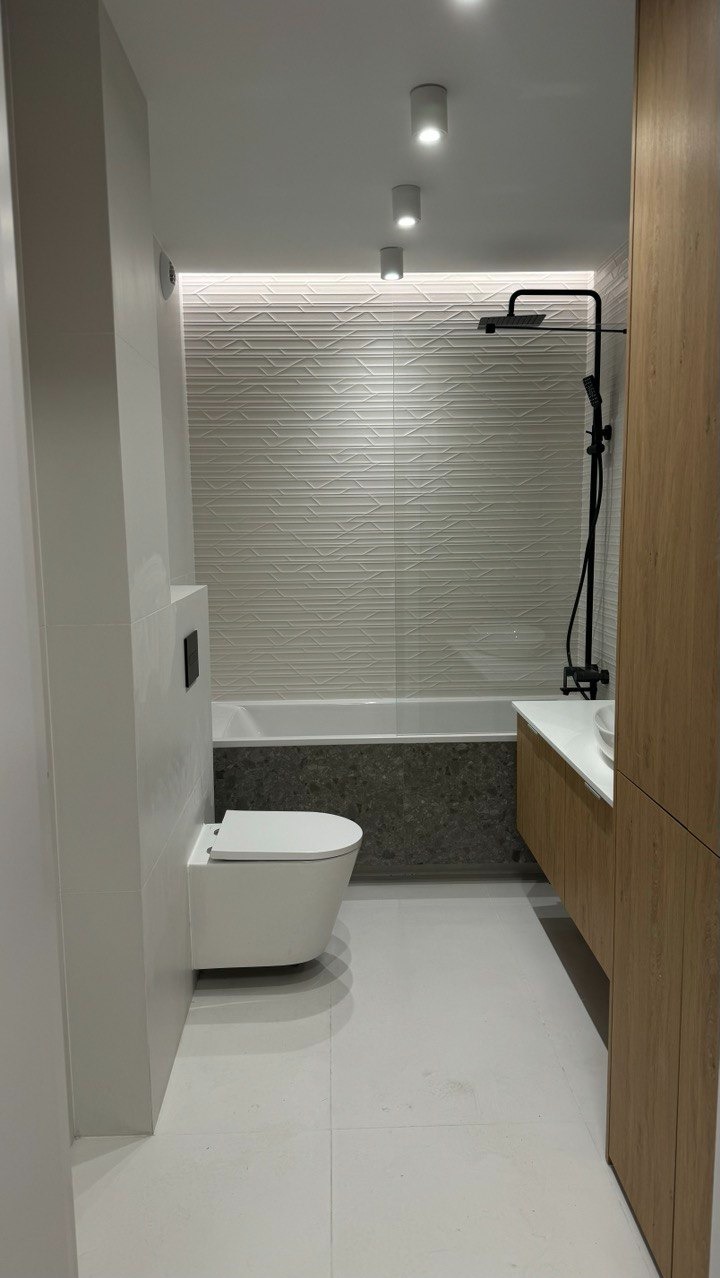 Nowoczesna łazienka z podwieszaną toaletą, białą armaturą, kabiną prysznicową typu walk-in z czarną baterią i drewnianą szafką pod umywalką, oświetlona punktowymi lampami sufitowymi i listwą LED...