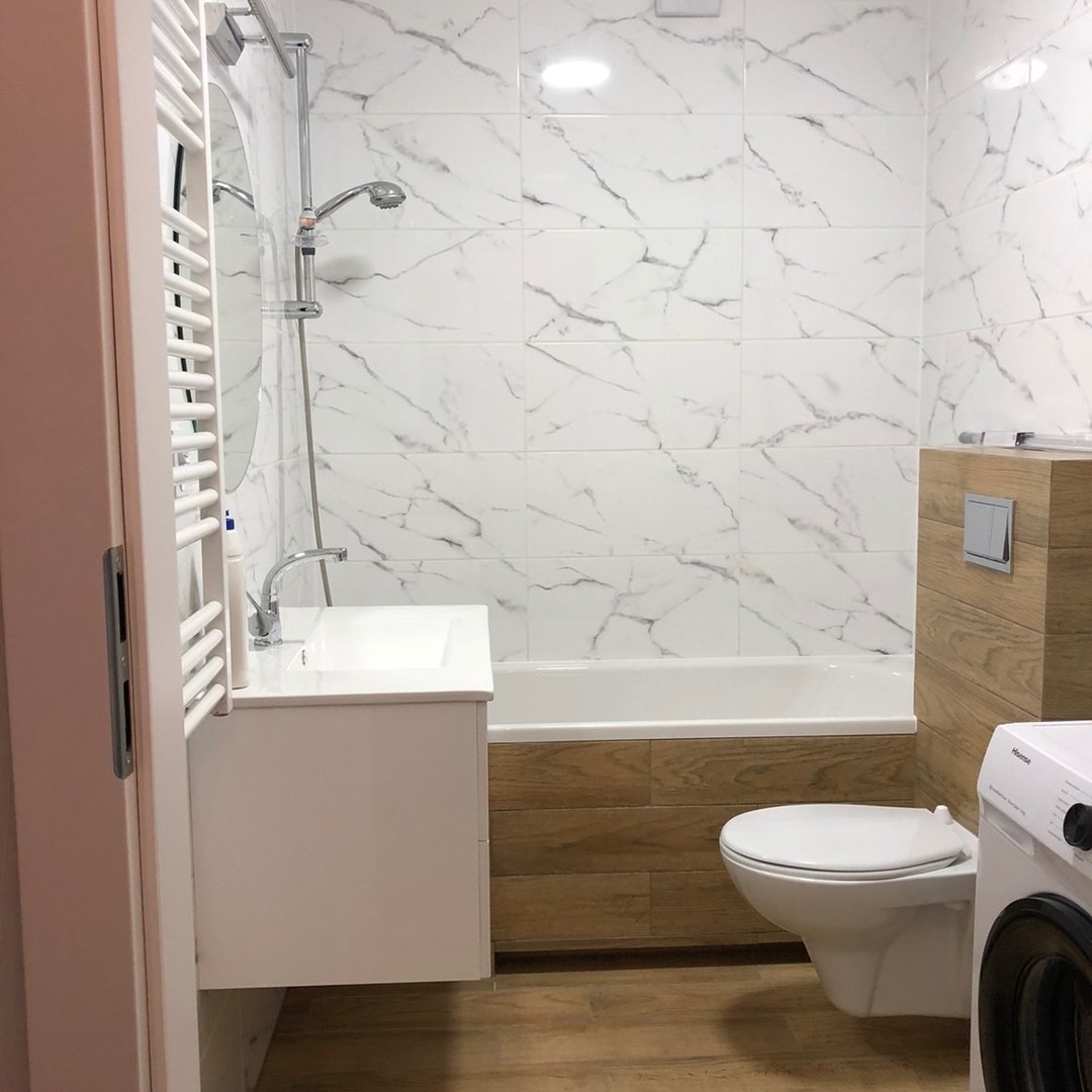 Nowoczesna łazienka po remoncie: białe płytki imitujące marmur, drewnopodobna obudowa wanny i toalety, biała umywalka z szafką oraz pralka w rogu.