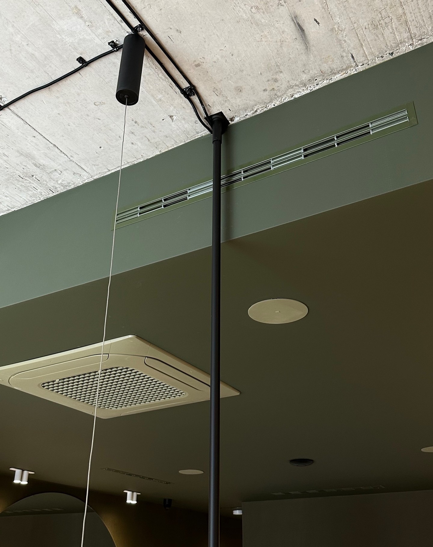 Szaro-zielone wnętrze z widoczną instalacją elektryczną na betonowym suficie, kratką wentylacyjną, lampą zwisającą na kablu i klimatyzatorem kasetonowym.