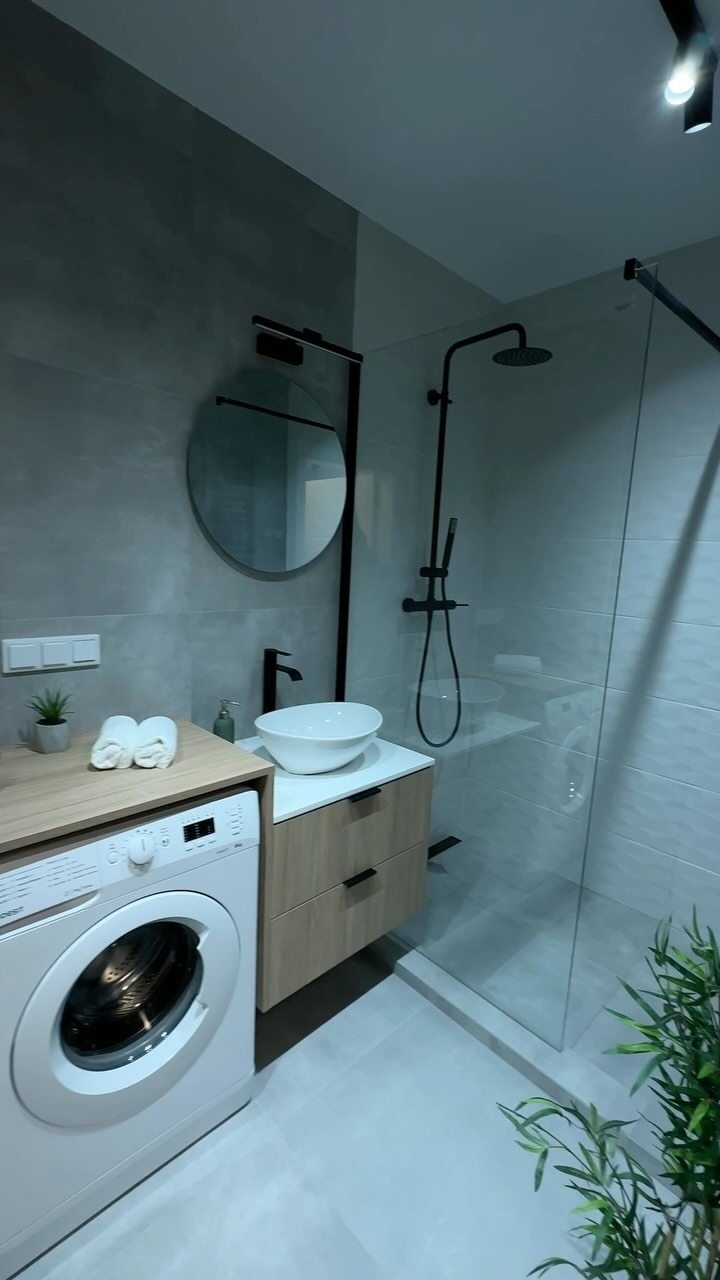 Nowoczesna łazienka z szarymi płytkami imitującymi beton, okrągłym lustrem z czarną ramą, umywalką nablatową, kabiną prysznicową typu walk-in z czarną armaturą oraz pralką pod drewnianym blatem...