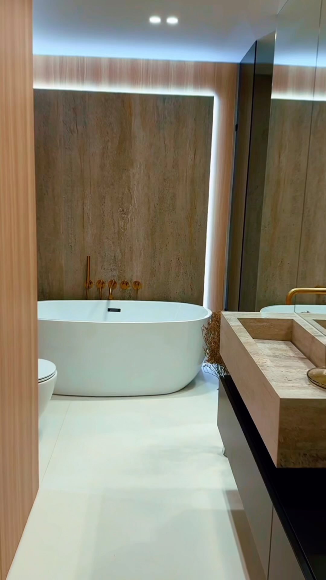 Nowoczesna łazienka z wolnostojącą wanną, armaturą w kolorze różowego złota i umywalką z kamienia, oświetlona ledowym paskiem za lustrem.