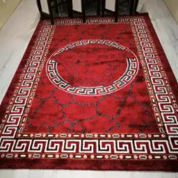 Czerwony, pluszowy dywan z motywem głowy Meduzy w stylu Versace, leżący na jasnej podłodze, widoczne krzesła w tle.